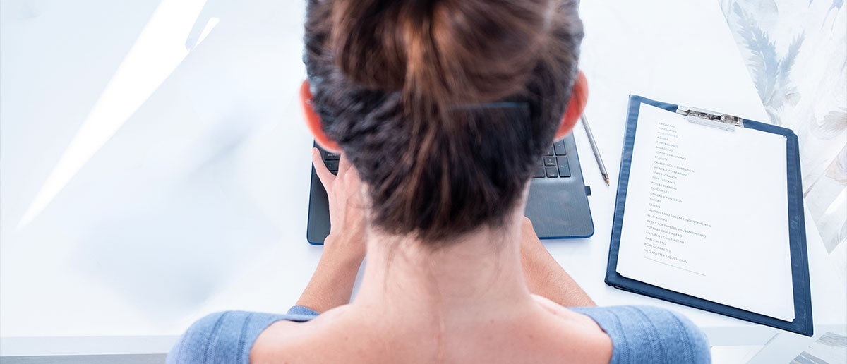 Primer plano de mujer de espaldas tecleando en un ordenador portail y un portapapeles con una lista a su derecha.